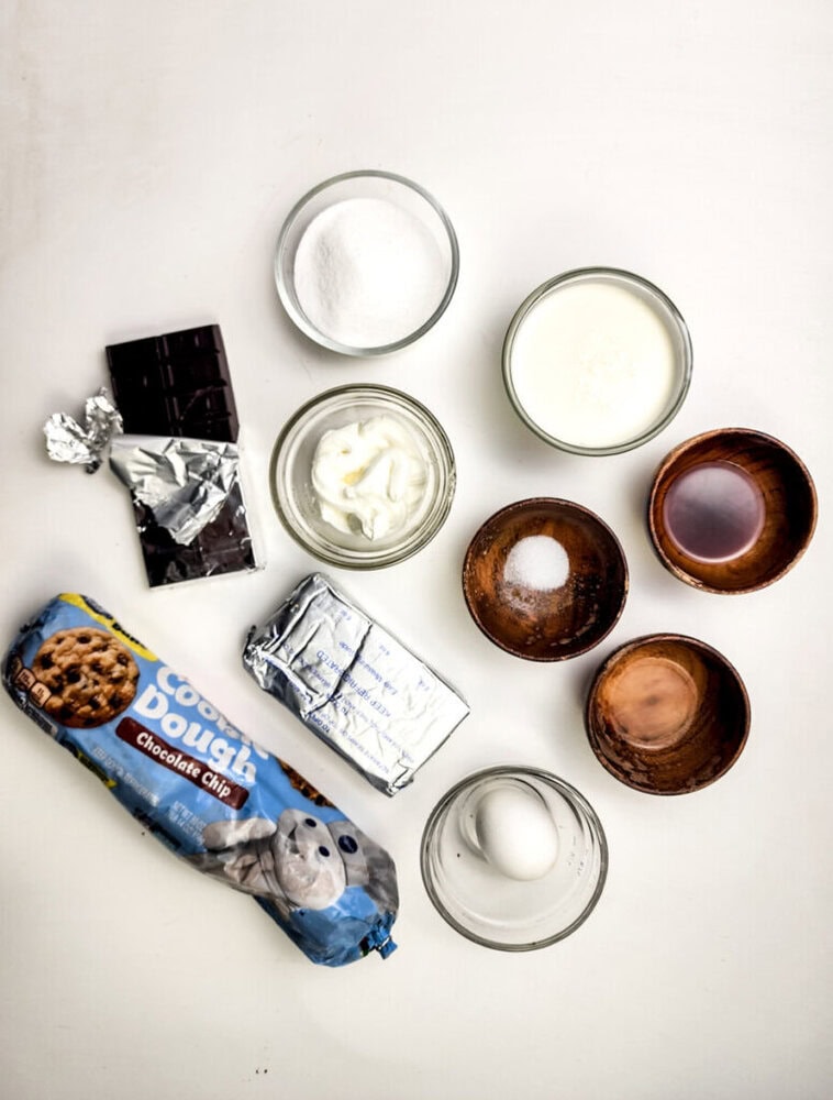 Assorted baking ingredients on a white surface, including chocolate, sugar, cream, vanilla, salt, butter, sour cream, an egg, and a package of chocolate chip cookie dough.