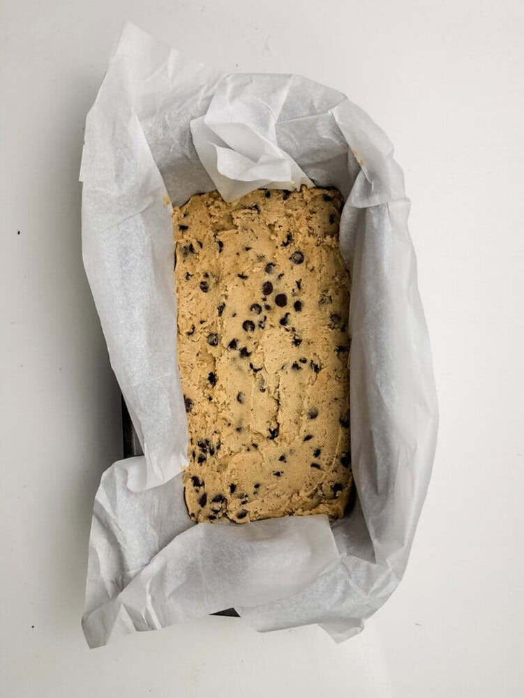 A loaf pan lined with parchment paper filled with unbaked chocolate chip cookie dough, viewed from above on a white surface.