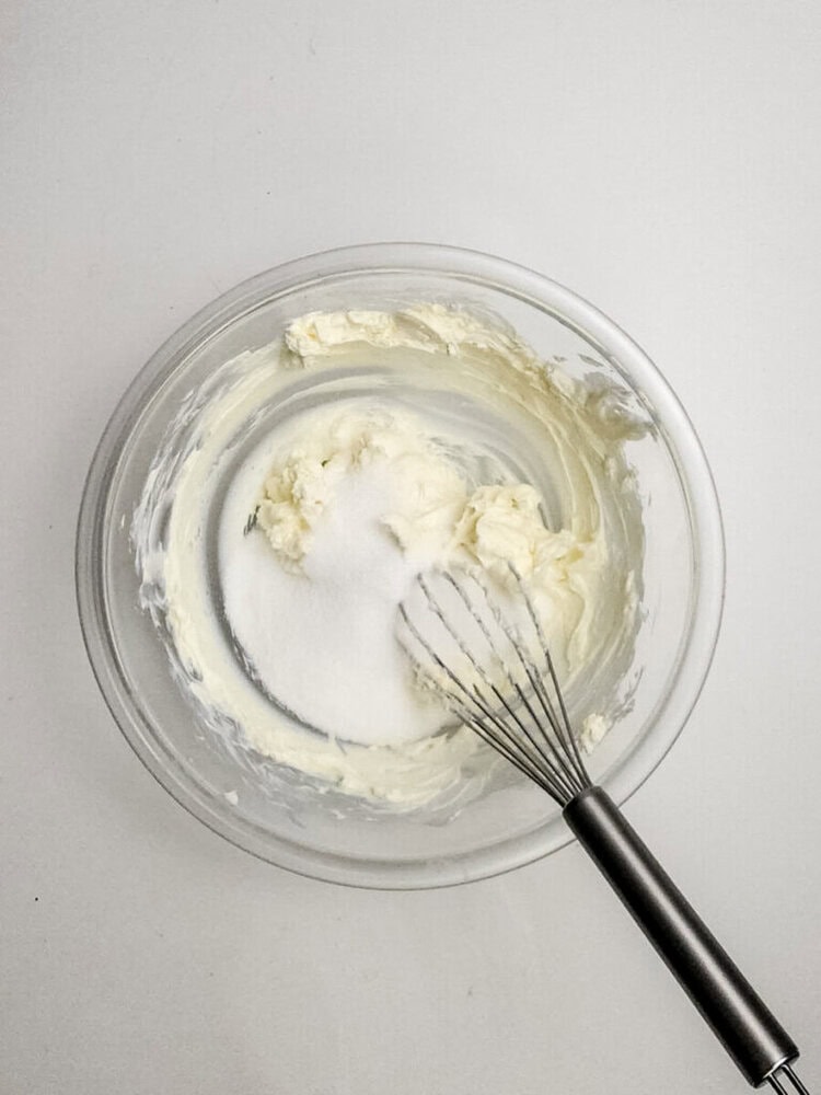 A glass bowl with creamed butter and sugar being mixed with a metal whisk, viewed from above on a white surface.