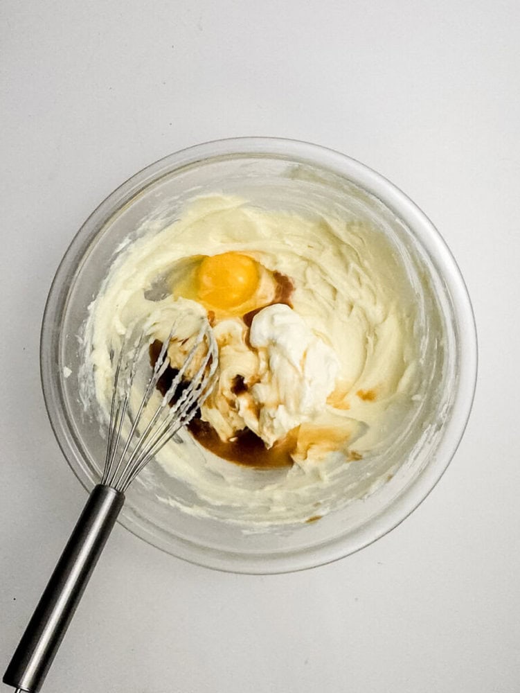 A glass bowl with creamed mixture, an egg, vanilla extract, and sour cream being mixed with a metal whisk on a white surface.