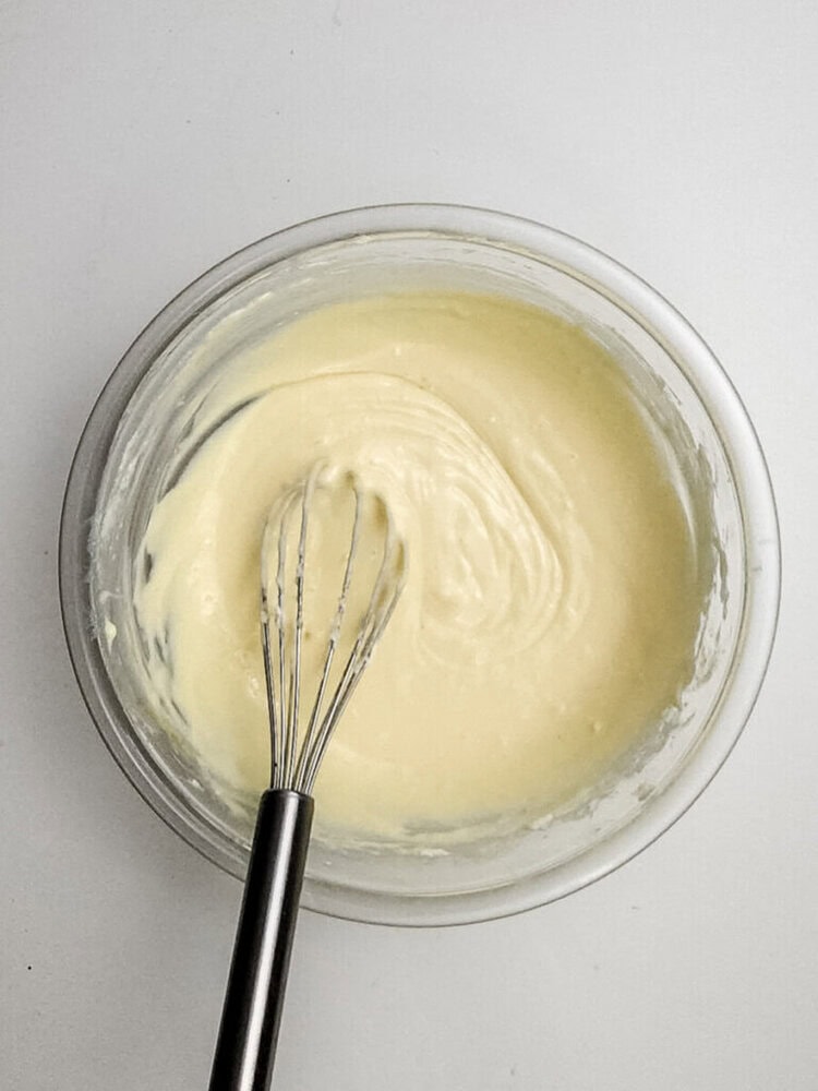 A metal whisk in a glass bowl of thick, pale yellow batter on a white surface.