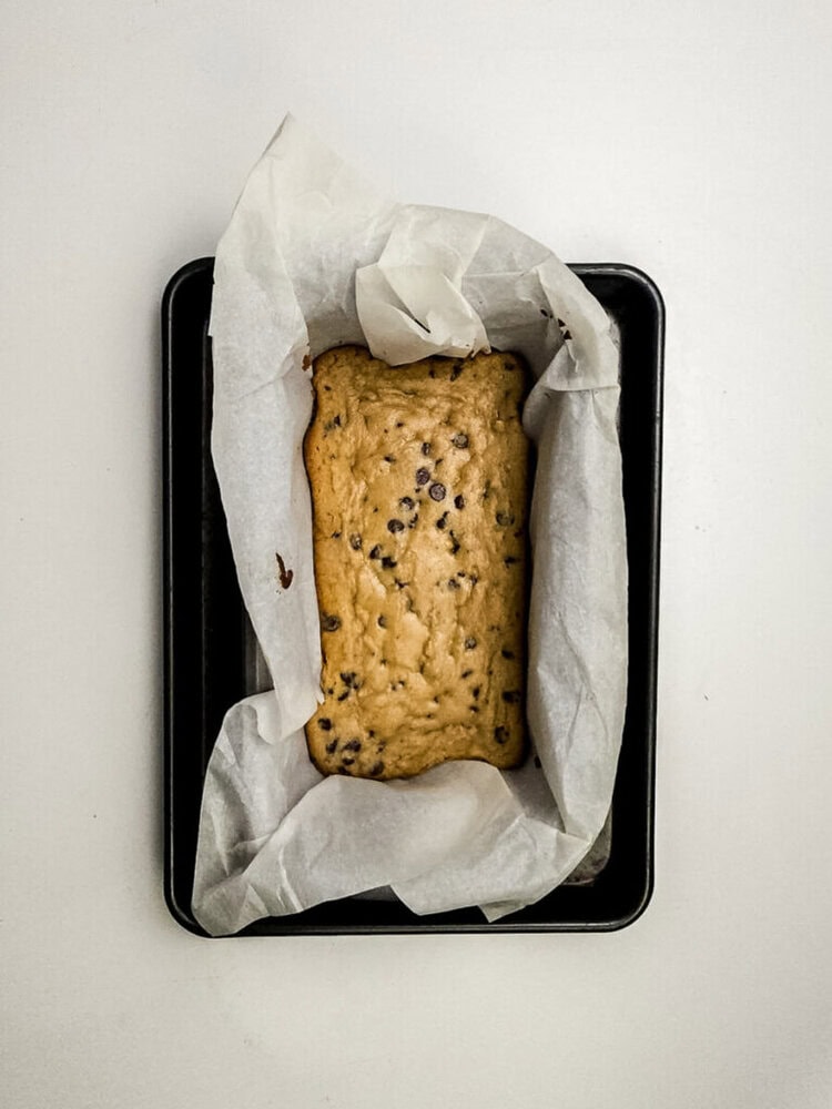 A loaf of chocolate chip banana bread sits on parchment paper in a black rectangular baking pan against a white background.