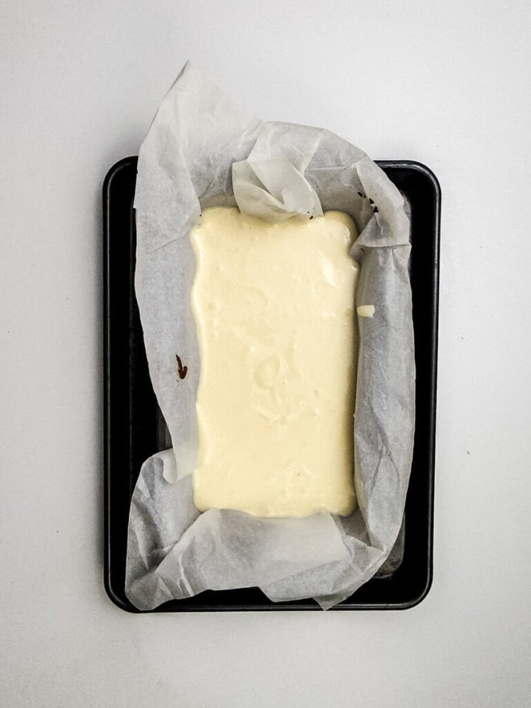 A rectangular baking tin lined with parchment paper holds a pale, unbaked batter on a white surface.