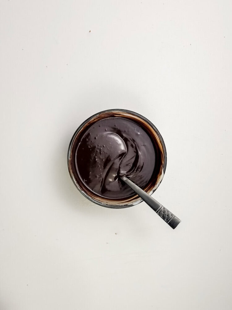 A bowl of smooth, glossy chocolate sauce with a metal spoon resting inside, set on a plain white surface.