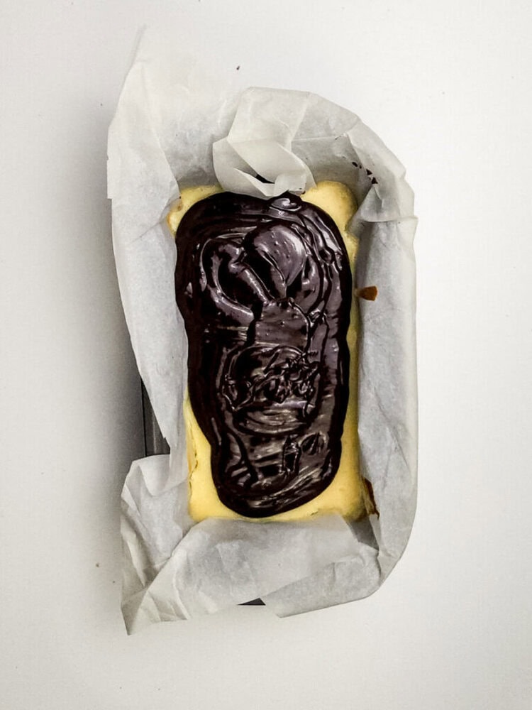 A rectangular loaf pan lined with parchment paper holds a baked cheesecake topped with a thick layer of glossy chocolate ganache.