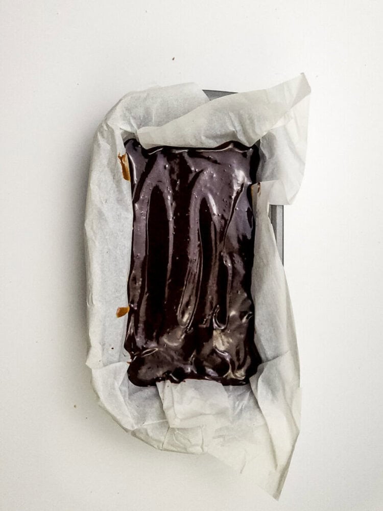 A rectangular pan lined with parchment paper holds a loaf covered in glossy chocolate glaze, viewed from above on a white surface.