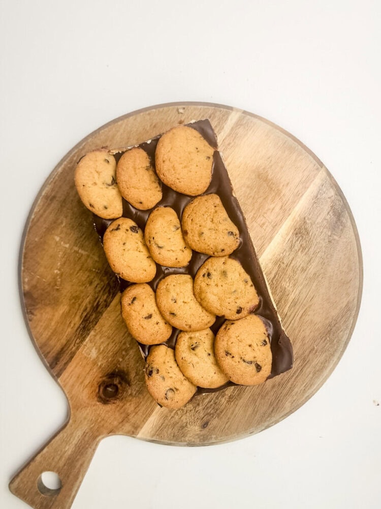 A rectangular chocolate dessert topped with chocolate chip cookies is placed on a round wooden cutting board.