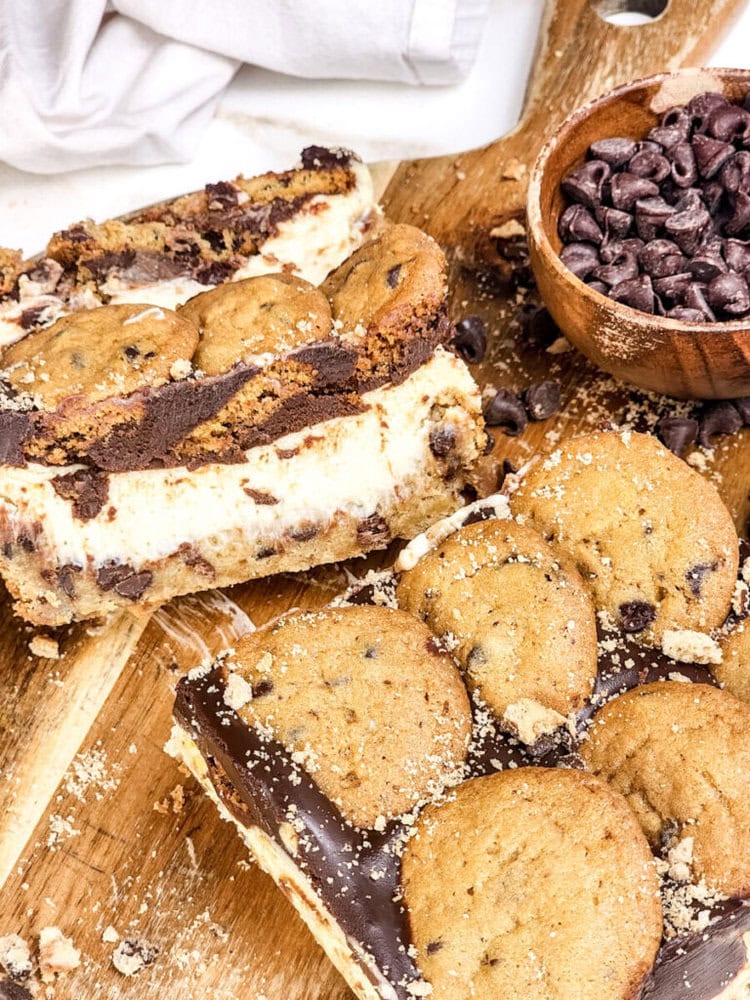 Chocolate chip cookie cheesecake bars and whole cookies displayed on a wooden board with a bowl of chocolate chips nearby.