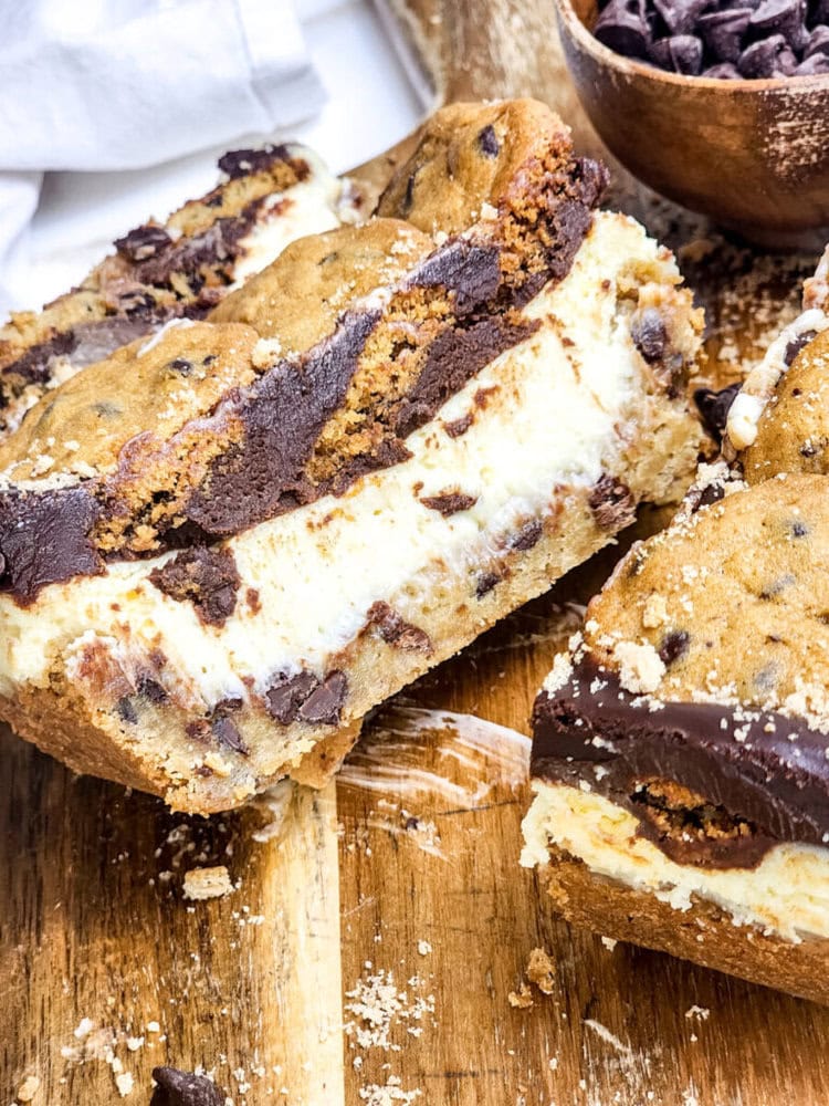 A close-up of a dessert with layers of chocolate chip cookie, cheesecake filling, and a chocolate topping, sliced on a wooden board.