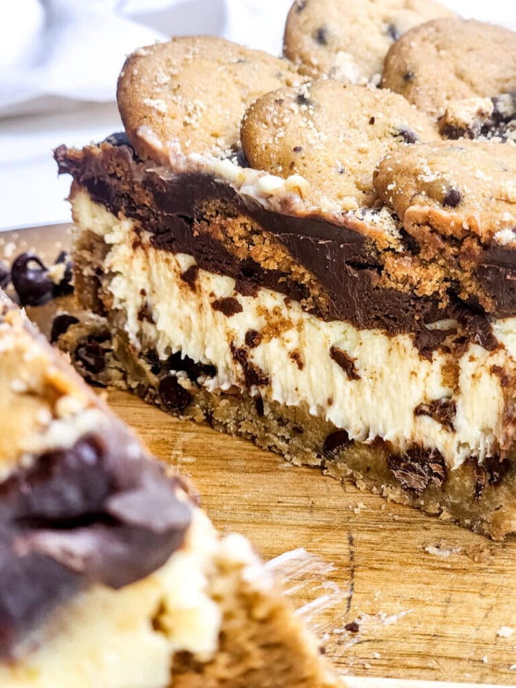 A close-up of a sliced layered dessert with chocolate chip cookie crust, creamy cheesecake filling, chocolate topping, and whole chocolate chip cookies on top.