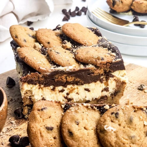 A layered dessert with chocolate chip cookies, creamy filling, and chocolate topping, displayed on a wooden board with extra cookies and chocolate chips scattered around.