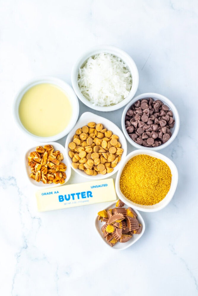 Seven small bowls and a stick of butter on a white surface, containing sweetened condensed milk, shredded coconut, chocolate chips, butterscotch chips, pretzels, graham cracker crumbs, and mini peanut butter cups.