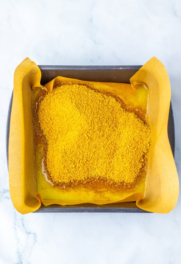 A baking pan lined with parchment paper, containing a layer of melted butter topped with a mound of yellow crumb mixture.