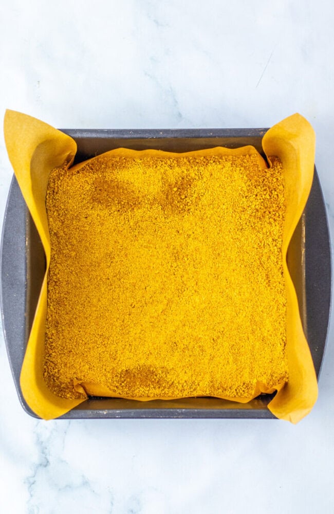 A rectangular baking pan lined with parchment paper, filled with a yellow, crumbly mixture, viewed from above on a white surface.