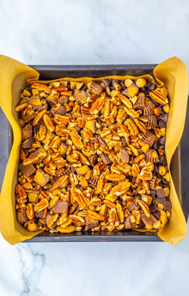 A square baking pan lined with parchment paper, filled with a mixture of chopped pecans, chocolate chips, and peanut butter cups.
