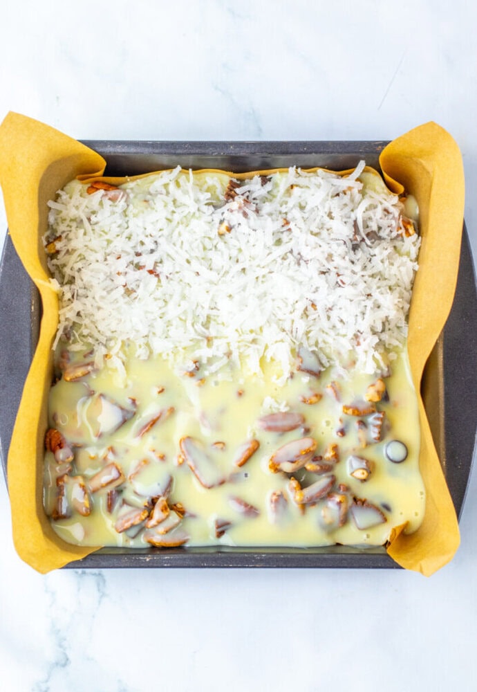 Square baking pan lined with parchment paper, filled halfway with coconut flakes and halfway with chopped nuts covered in sweetened condensed milk.
