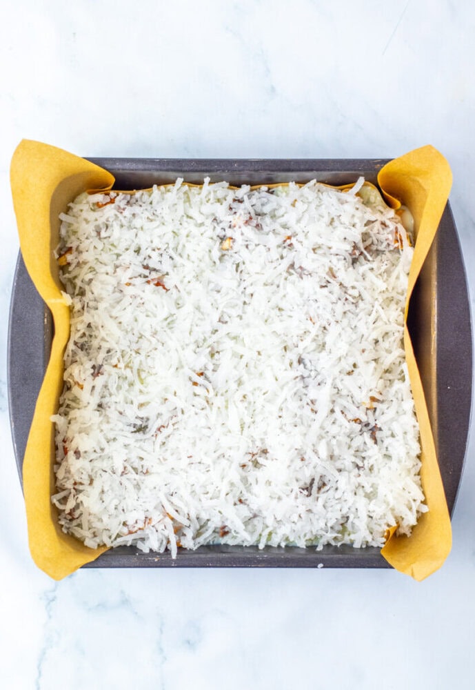 A square baking pan lined with parchment paper, filled with an even layer of shredded coconut mixture, ready to be baked.