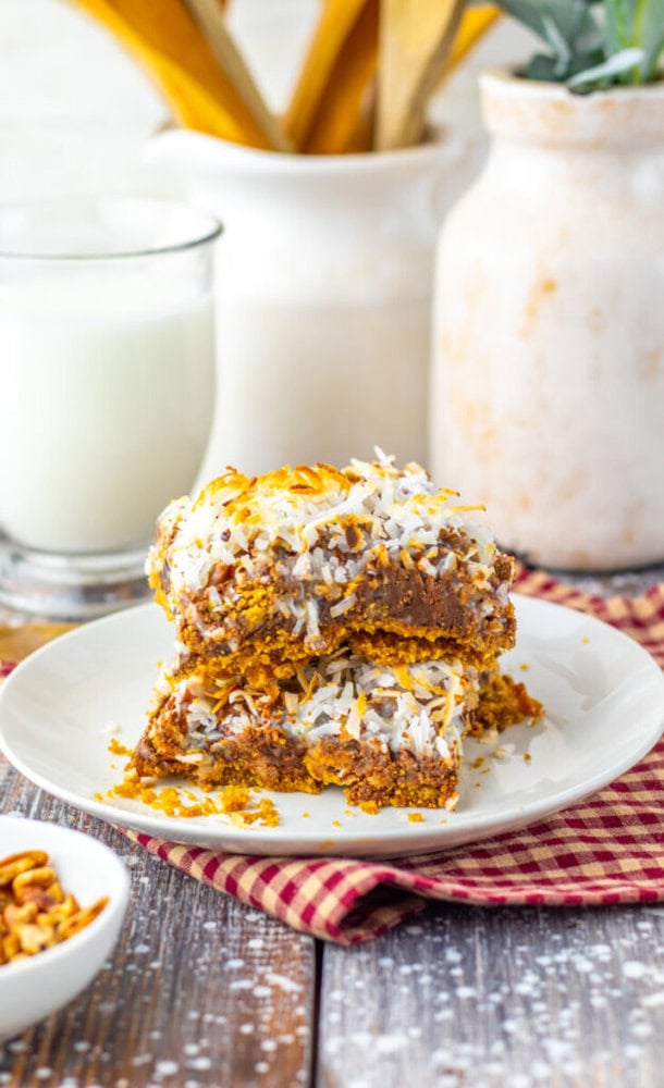 Two dessert bars topped with shredded coconut are stacked on a white plate, with a glass of milk, a small bowl of nuts, and kitchen items in the background.