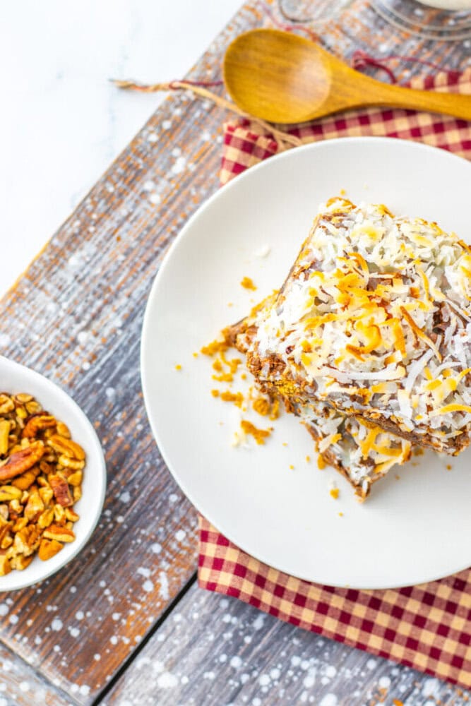 A square dessert topped with shredded coconut and orange zest on a white plate, next to a bowl of granola and a wooden spoon on a checked napkin.