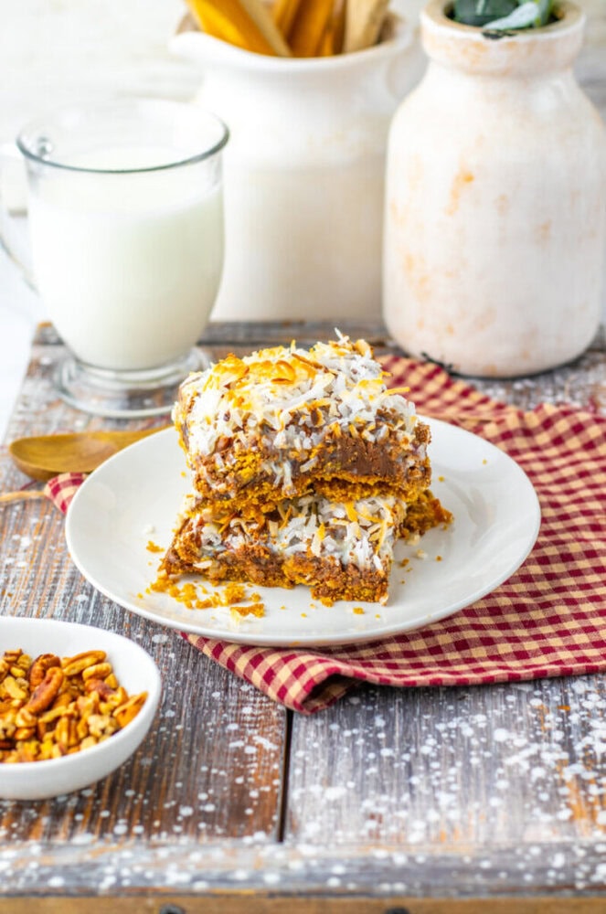 Two square dessert bars topped with shredded coconut are stacked on a white plate, set on a red checkered cloth, with a glass of milk and a small bowl of nuts nearby.