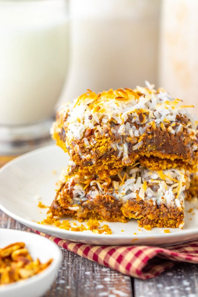 A close-up of two dessert bars topped with shredded coconut, stacked on a white plate, with a glass of milk in the background.