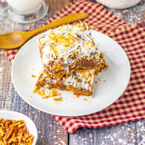 Two pieces of coconut and nut-topped dessert bars stacked on a white plate, placed on a red checkered cloth with a wooden spoon and a small bowl of pecans nearby.