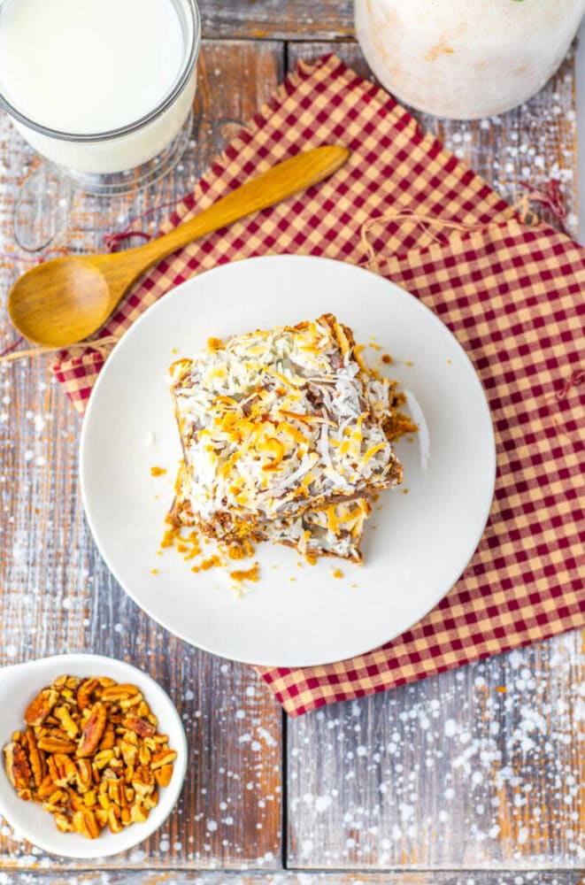 A square piece of cake topped with coconut and nuts on a white plate, set on a red checkered napkin with a wooden spoon, a small bowl of pecans, and a glass of milk nearby.