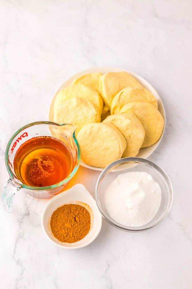 A plate of biscuit dough rounds, a measuring cup of honey, a bowl of white sugar, and a small dish of ground cinnamon on a white surface.