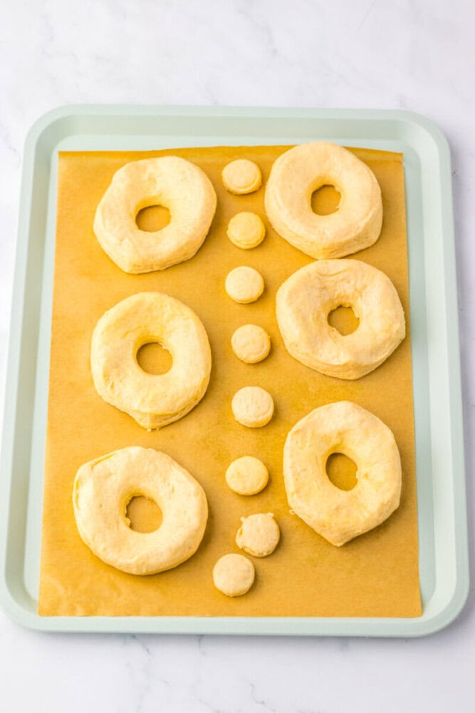Six unbaked doughnuts and six doughnut holes are arranged on a parchment-lined baking sheet.