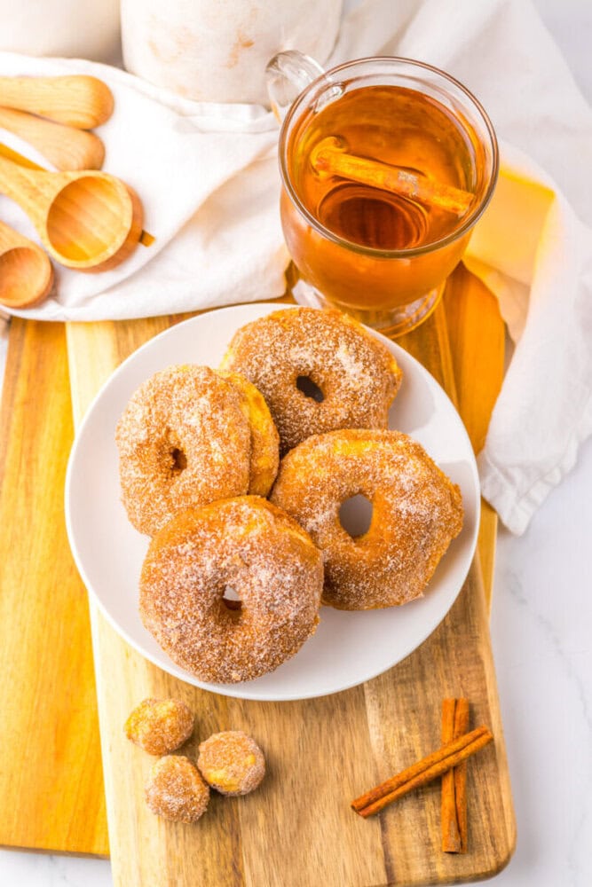 A white plate with sugar-coated donuts and donut holes on a wooden board, next to a glass mug of tea with a cinnamon stick and wooden measuring spoons.