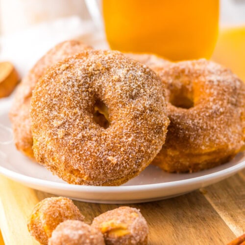 A plate of sugar-coated donuts sits on a wooden board, with small donut holes and cinnamon sticks nearby.