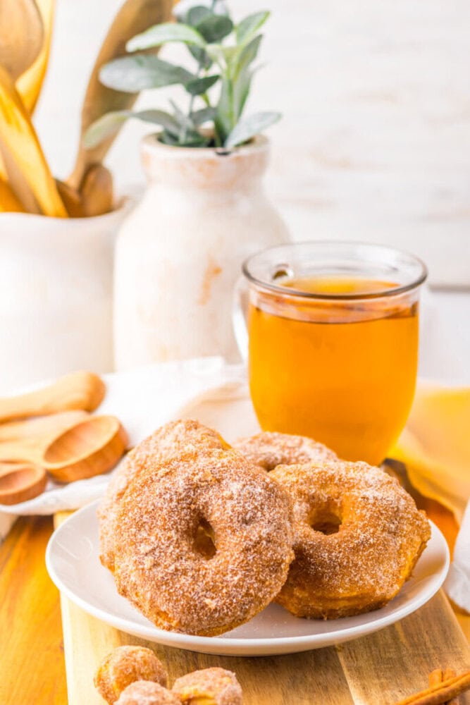 A plate of sugar-coated donuts is in front of a glass mug filled with tea; kitchen utensils and a potted plant are in the background.