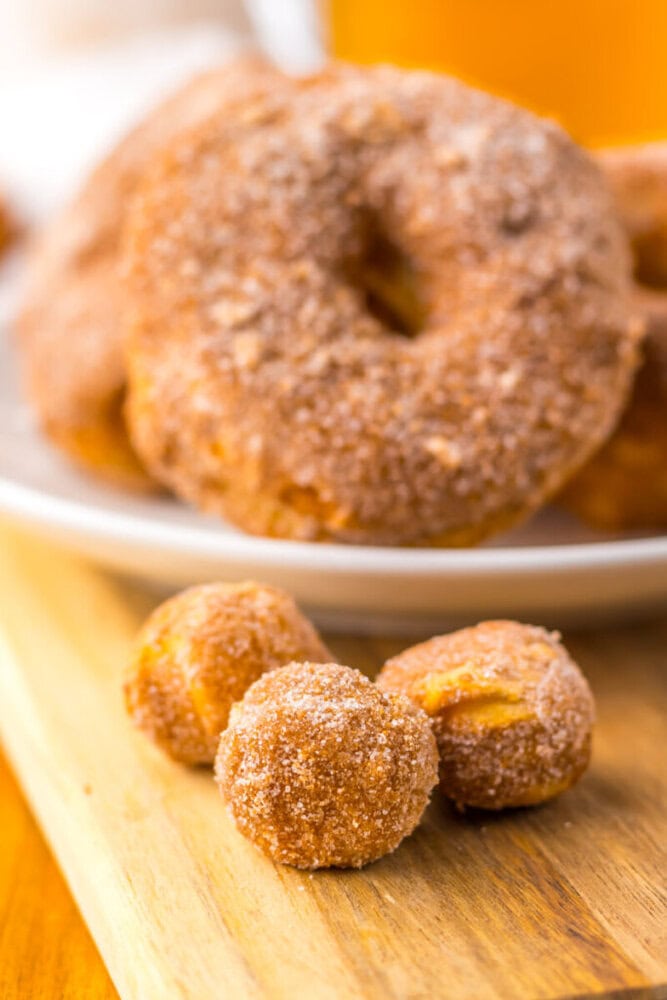 Three cinnamon sugar-coated donut holes are on a wooden board, with a plate of full-sized donuts in the blurred background.