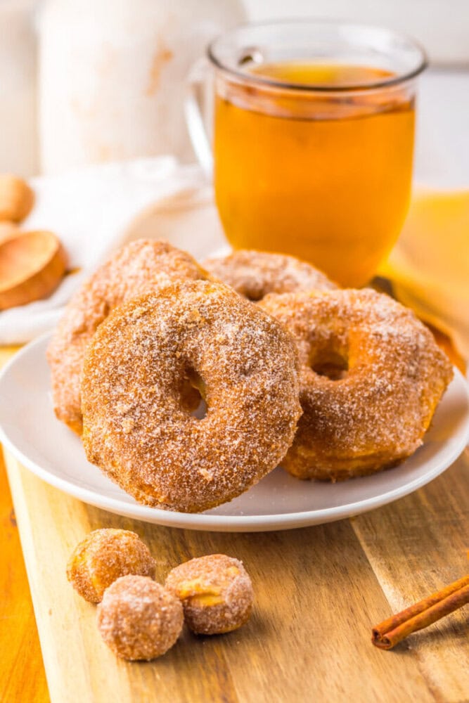 A plate of sugar-coated donuts and donut holes sits on a wooden board beside a clear glass mug of tea.