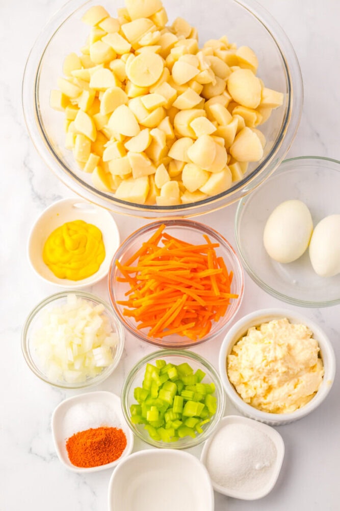 A variety of ingredients in bowls, including chopped potatoes, hard-boiled eggs, carrots, celery, onion, mayonnaise, mustard, seasoning, and sugar, arranged on a white surface.