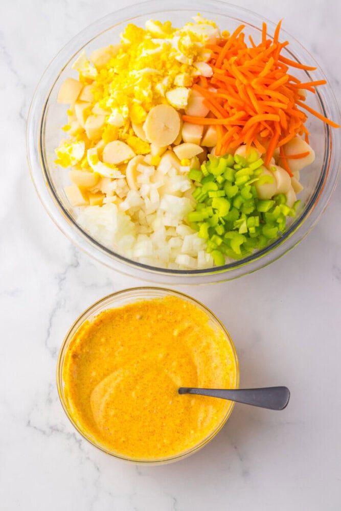 A glass bowl with chopped boiled eggs, carrots, celery, onions, and hearts of palm; next to it is a bowl of yellow dressing with a spoon.