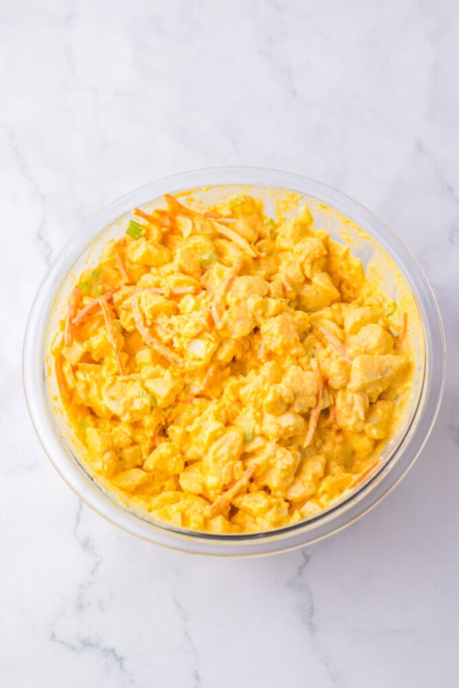 A glass bowl filled with a yellow, creamy cauliflower salad mixed with sliced carrots and green vegetables, placed on a white marble surface.