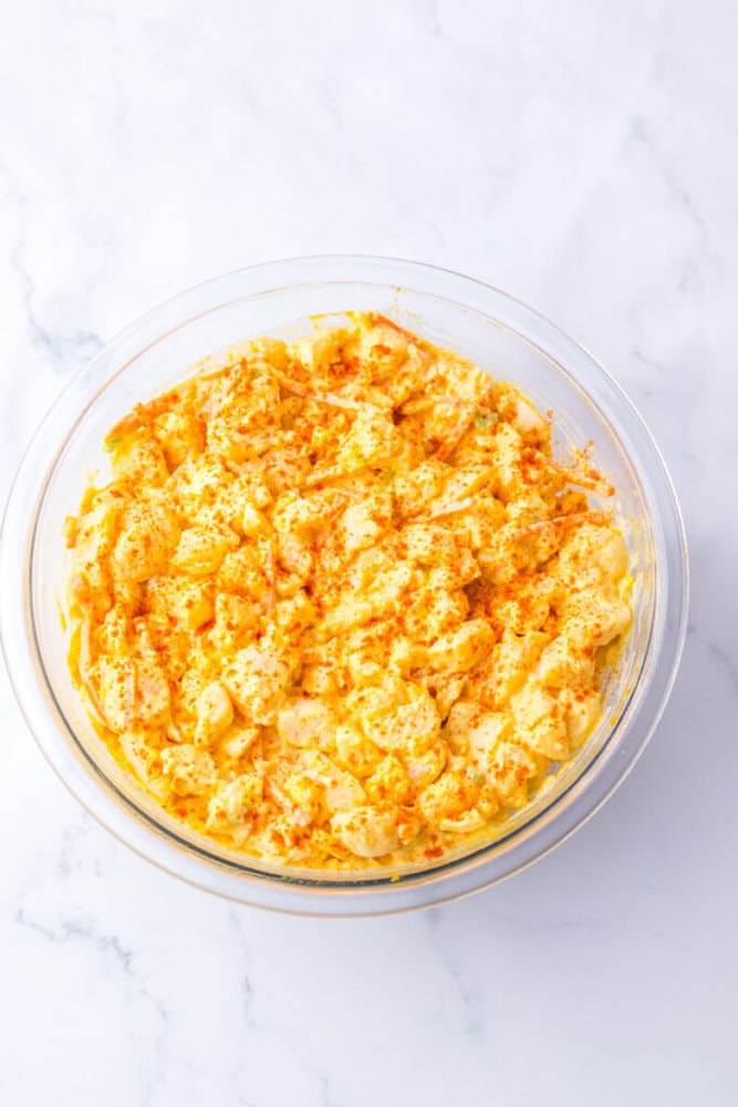 A glass bowl filled with deviled egg potato salad, topped with a generous sprinkle of paprika, on a white marble surface.