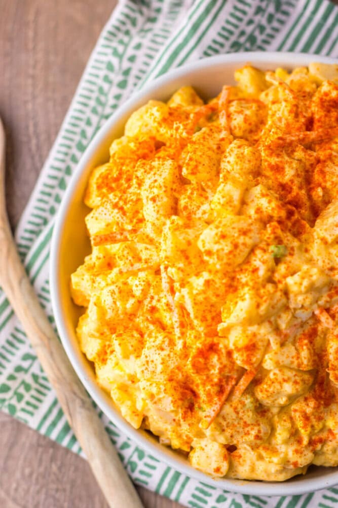 A bowl of macaroni salad topped with paprika sits on a green and white patterned cloth next to a wooden spoon.