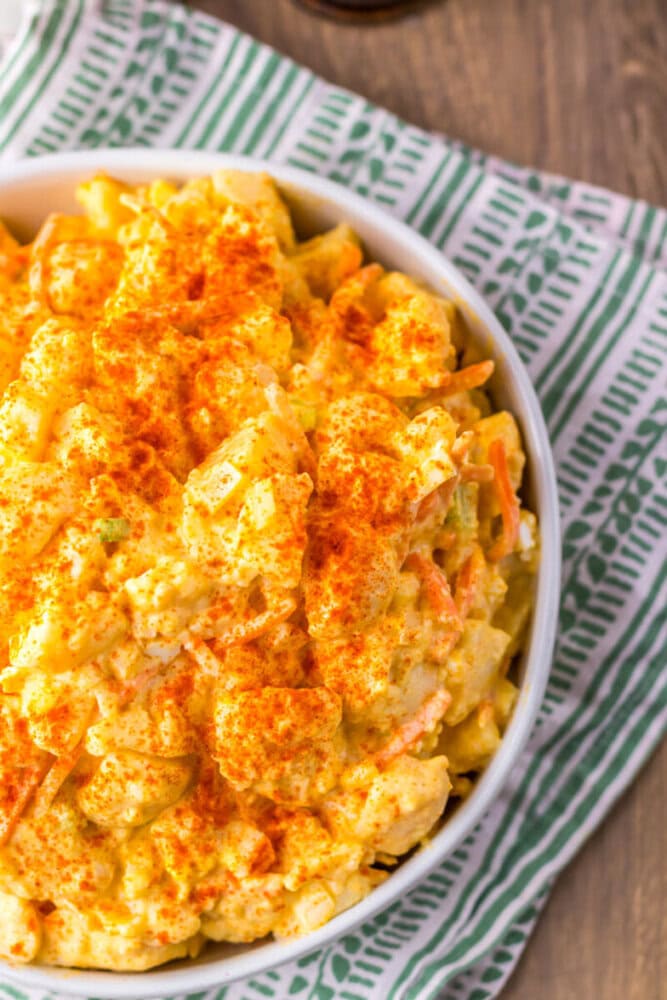 A bowl of potato salad topped with paprika sits on a patterned green and white cloth on a wooden surface.