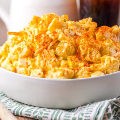 A white bowl filled with creamy macaroni and cheese, topped with a sprinkle of paprika, sits on a green and white striped cloth next to a wooden spoon.