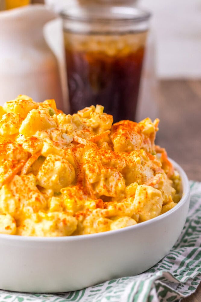 A white bowl filled with creamy potato salad topped with paprika, sitting on a striped cloth with a drink and condiment bottles in the background.