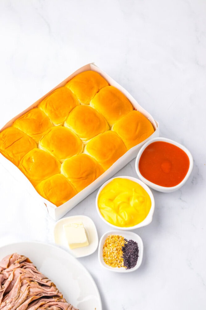 A tray of twelve golden dinner rolls next to small bowls of mustard, red sauce, butter, seasonings, and a plate of sliced roast beef on a white surface.