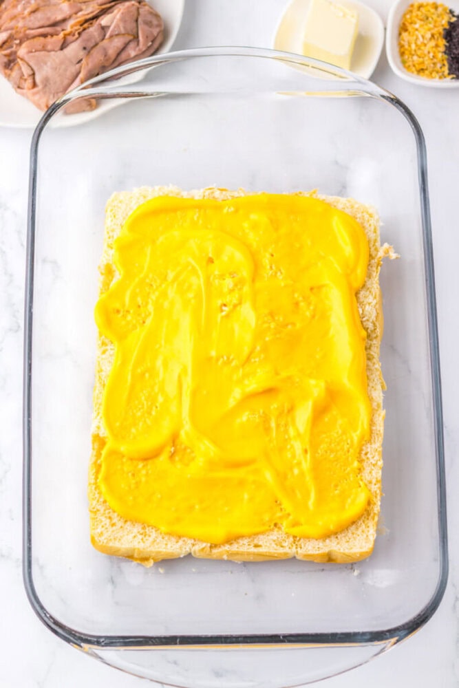Glass baking dish with a large slice of bread spread with yellow mustard, surrounded by bowls of roast beef, butter, and seasoning on a white surface.