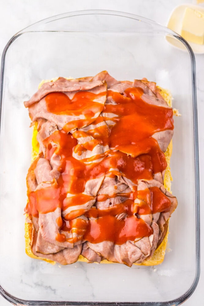 Slices of roast beef topped with barbecue sauce are layered on a bottom half of bread in a glass baking dish.
