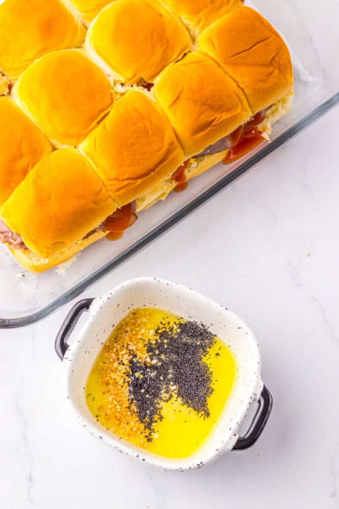 A baking dish with sandwiches next to a small bowl containing melted butter, minced garlic, dried onion, and poppy seeds on a white surface.