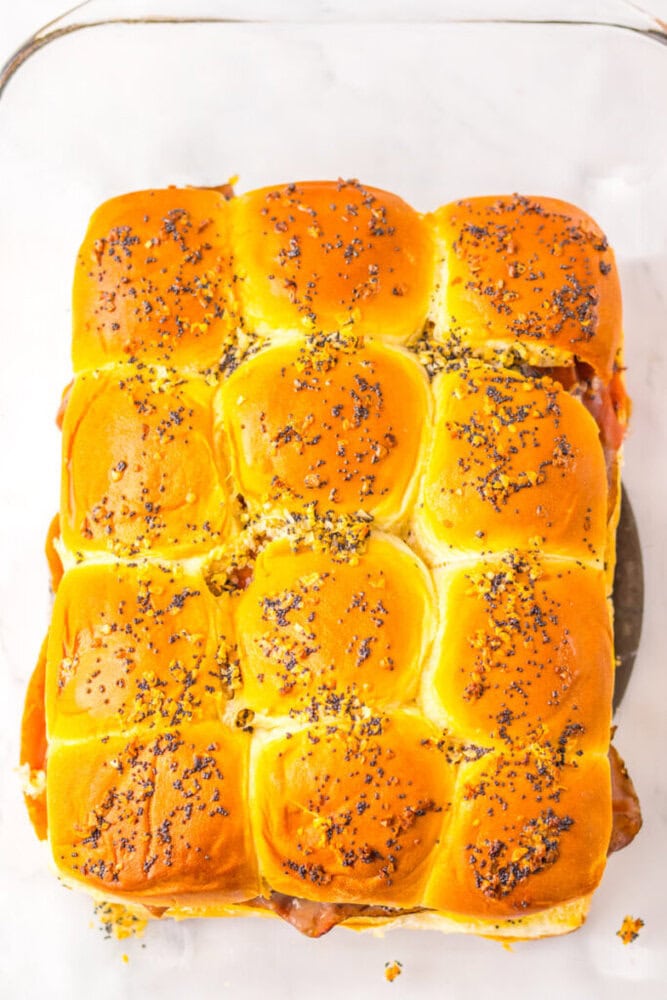 A tray of twelve baked sandwich sliders topped with poppy seeds, arranged closely together on a glass baking dish.