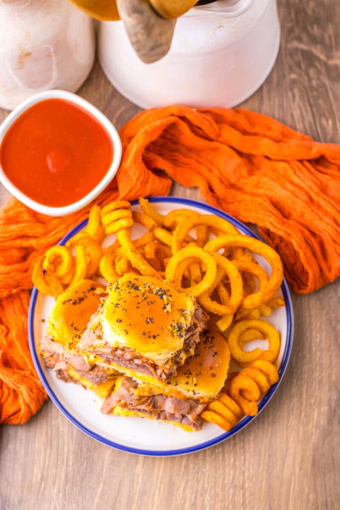 A plate with sliders topped with cheese and poppy seeds, served with curly fries. A bowl of red dipping sauce and orange cloth are nearby on a wooden surface.