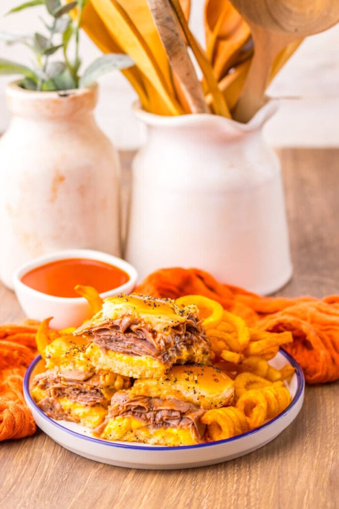 A plate with a stacked cheesy beef sandwich and curly fries, with a bowl of dipping sauce and kitchen utensils in the background.