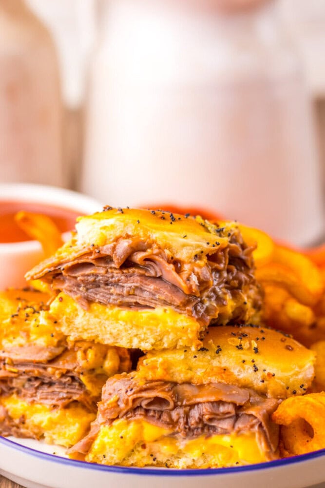 Close-up of stacked roast beef and cheese sliders on poppy seed buns, served with curly fries and a small dish of dipping sauce.